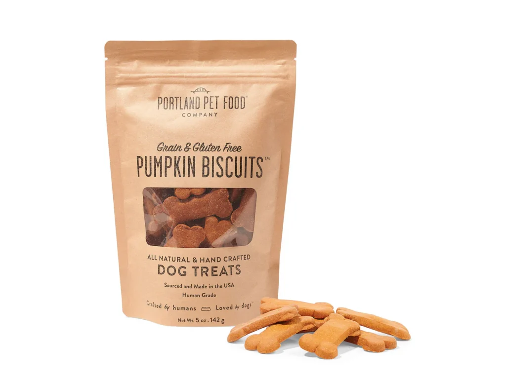 A 5 oz brown paper bag of Portland Pet Food Company Pumpkin Biscuits featuring a clear window showing bone-shaped treats, with several loose biscuits scattered on the white surface in front of the bag.